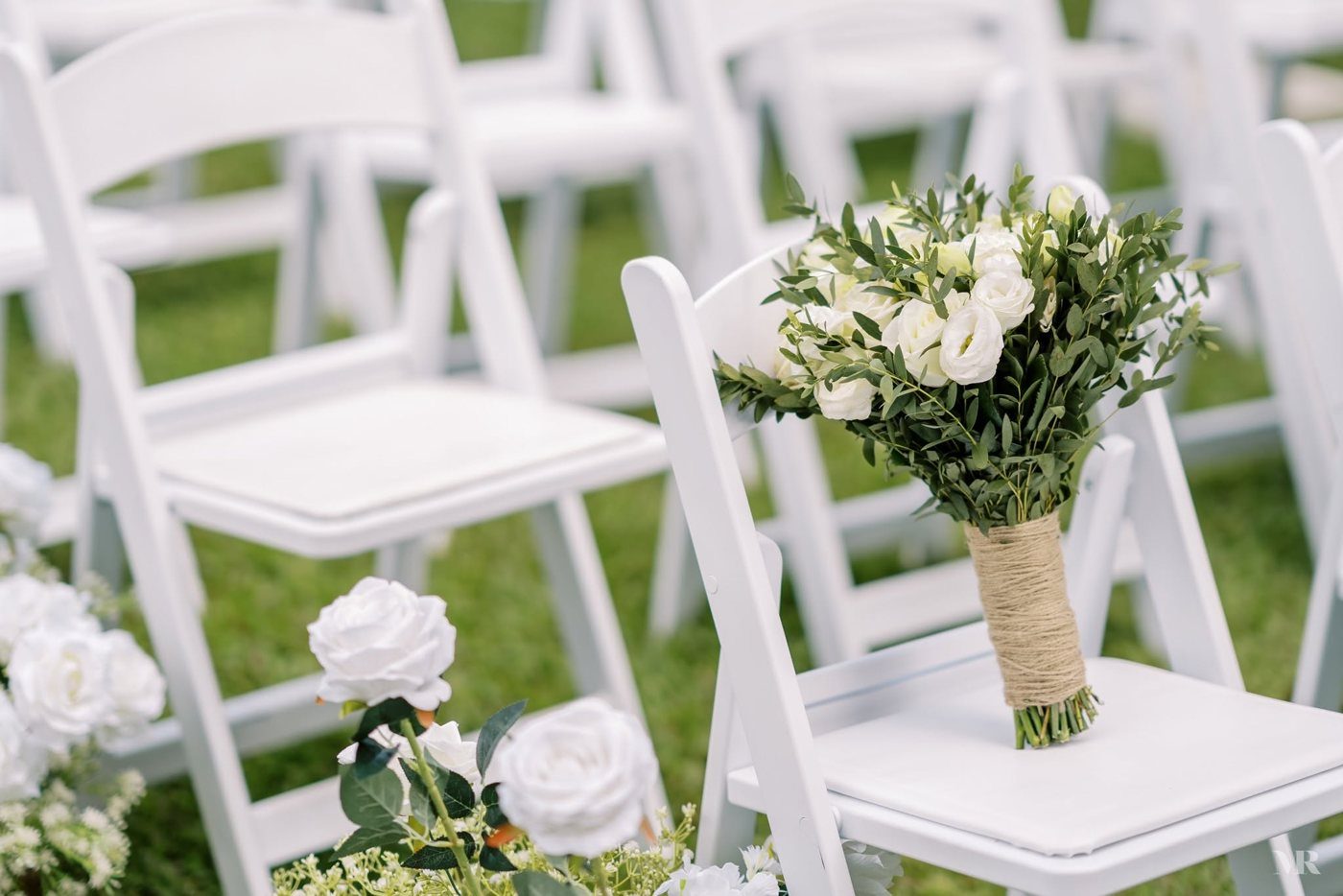 White ceremony chairs on the grass with a bouquet of white roses and greenery